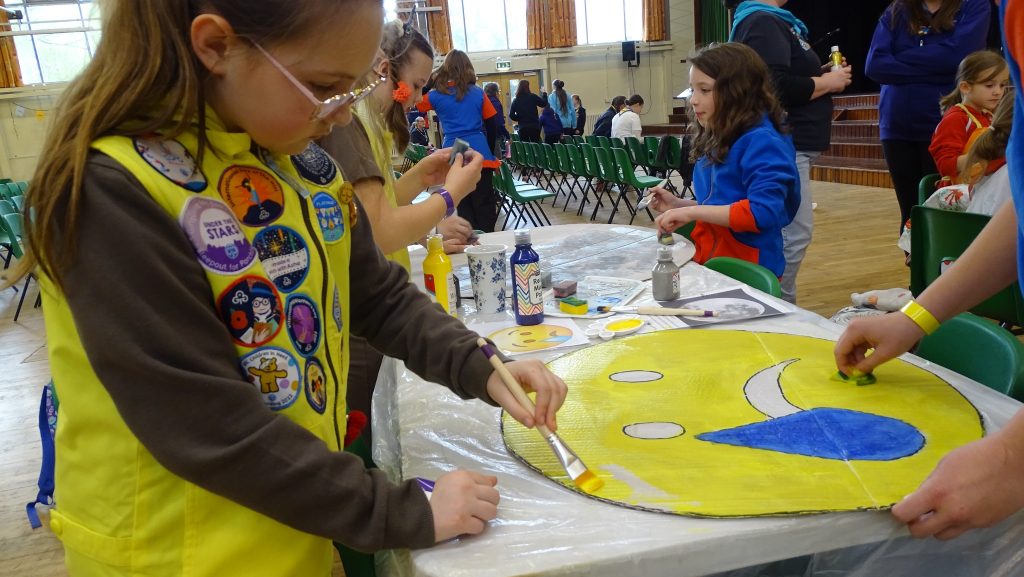 A Brownie painting a face pace board to hold up, with Guides in the background.
